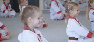Junge Taekwondo-Schüler in weißen Anzügen mit rotem Kragen sitzen im Schneidersitz auf dem Dojo-Boden und hören aufmerksam dem Unterricht zu.