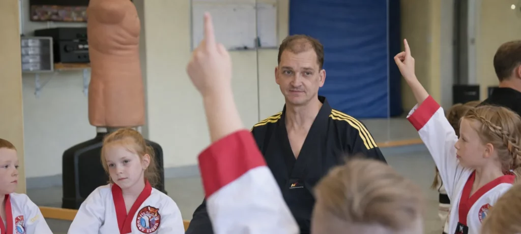 Taekwondo-Trainer im schwarzen Anzug unterrichtet Kinder, die im Dojo sitzen und ihre Hände heben.