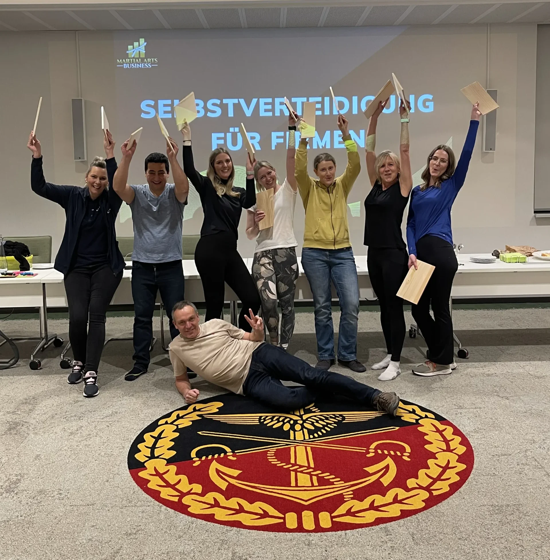 Workshop-Gruppe mit erhobenen Armen und Trainingsbrettern vor der Präsentation Selbstverteidigung für Firmen